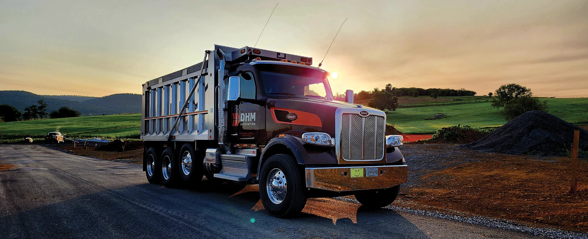 DHM dump truck driving along road with sun shining behind the truck.
