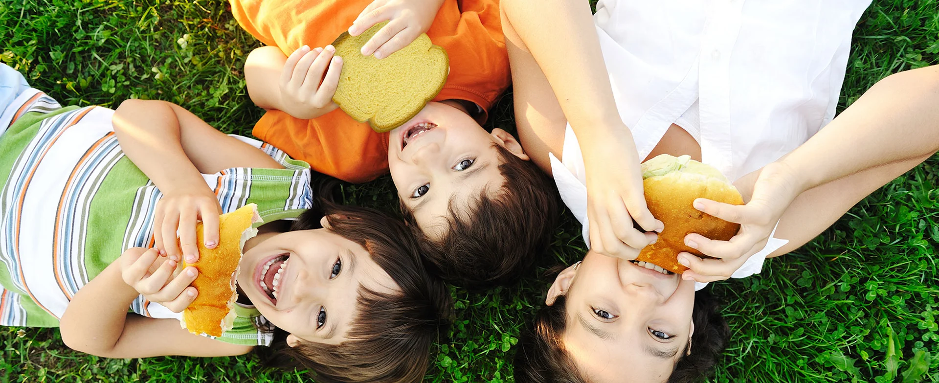 Smiling children laying down in grass eating potato rolls and bread