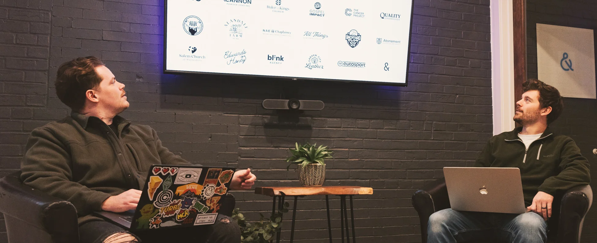 Branding & Identity meeting. Two employees, Dylan and Adam, are on their laptops and looking at a central screen.