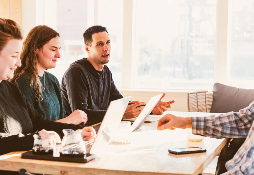 Four individuals are seated at a wooden table, engaged in a discussion with laptops and glasses in front of them. Sunlight streams in from a window, illuminating the scene.