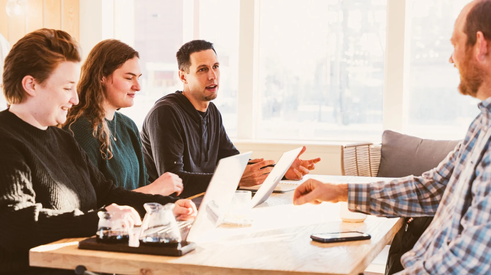 Four individuals are seated at a wooden table, engaged in a discussion with laptops and glasses in front of them. Sunlight streams in from a window, illuminating the scene.