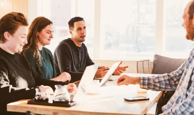 Four individuals are seated at a wooden table, engaged in a discussion with laptops and glasses in front of them. Sunlight streams in from a window, illuminating the scene.