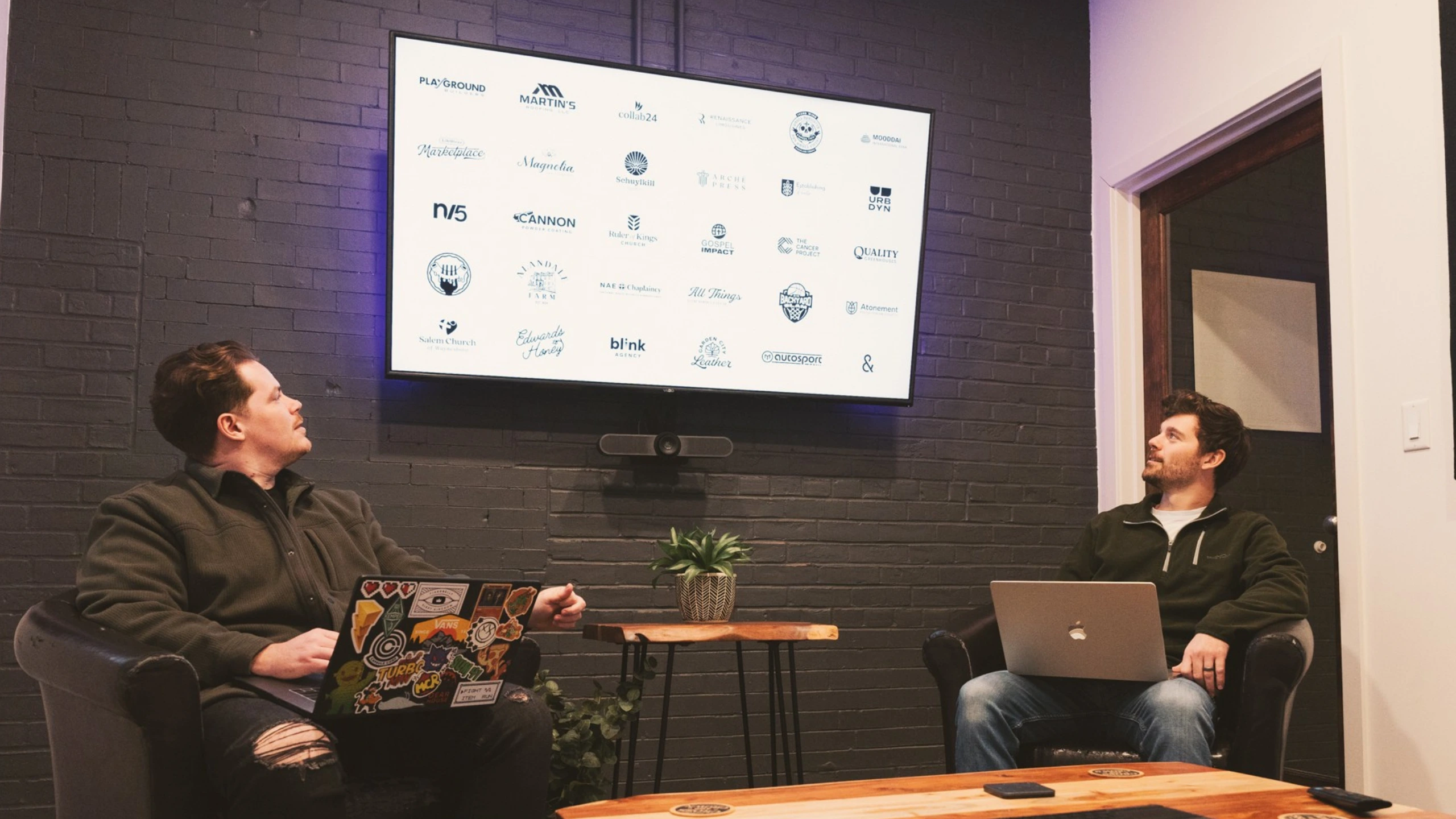 Two people are sitting in chairs in front of a large screen displaying various logos. One person has a laptop on their lap, and the other has a laptop on their lap as well, with a tablet on the coffee table in front of them.