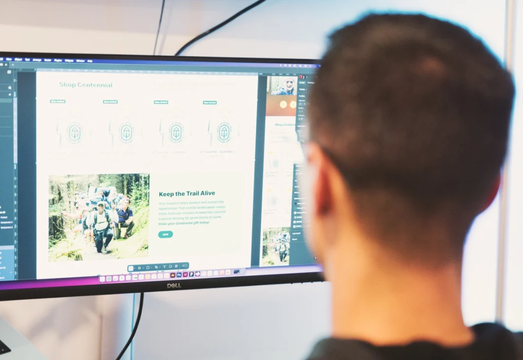 A person looks at a computer screen displaying a website with images of people hiking and text about graphics