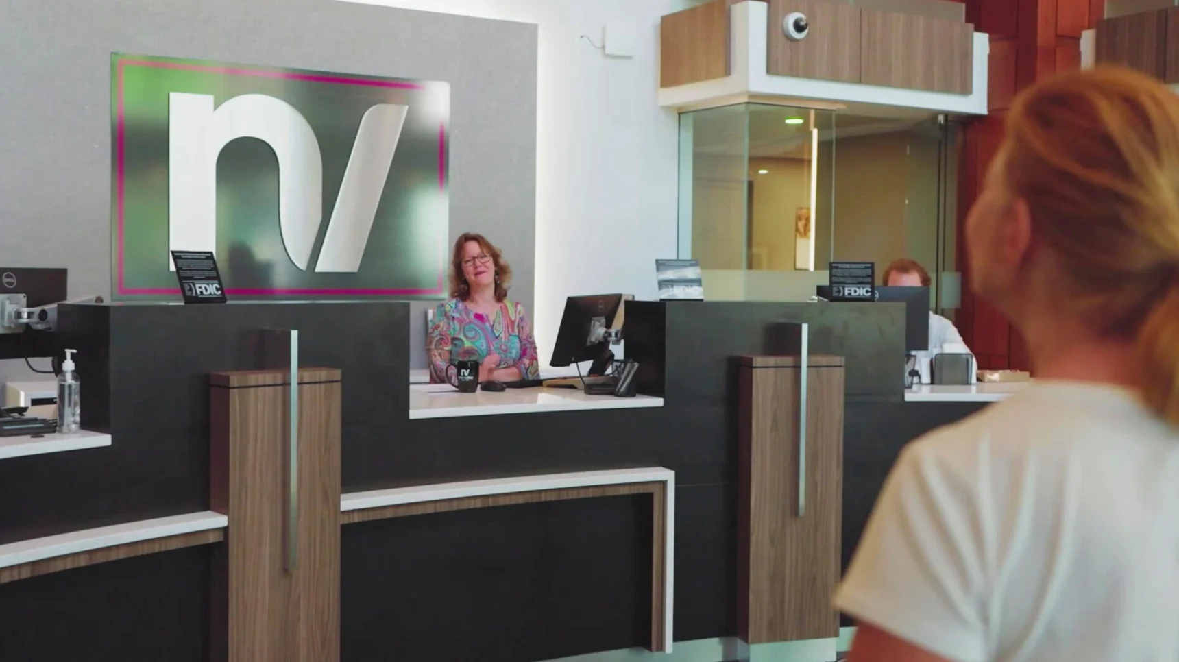 A woman with glasses and a colorful shirt smiles from behind a reception desk with a large logo. Another person is visible behind a glass partition at the desk.
