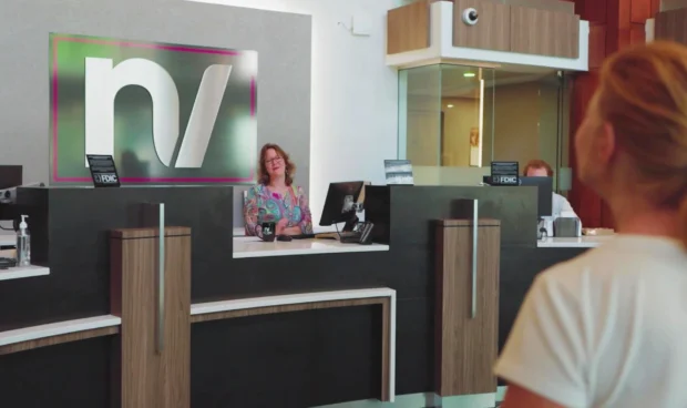 A woman with glasses and a colorful shirt smiles from behind a reception desk with a large logo. Another person is visible behind a glass partition at the desk.