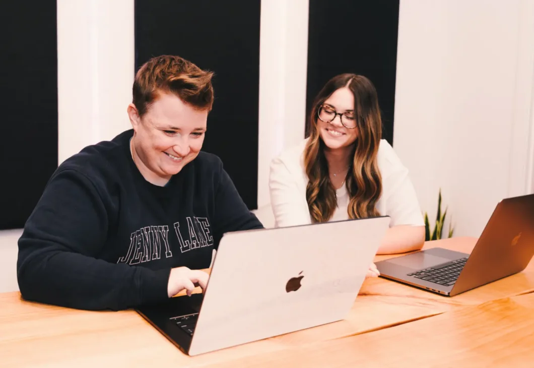 Two individuals are seated at a wooden table, each with a laptop in front of them. The person on the left is wearing a navy sweatshirt and smiling while looking at their laptop. The person on the right is wearing a white shirt and glasses, also looking at their laptop and smiling.