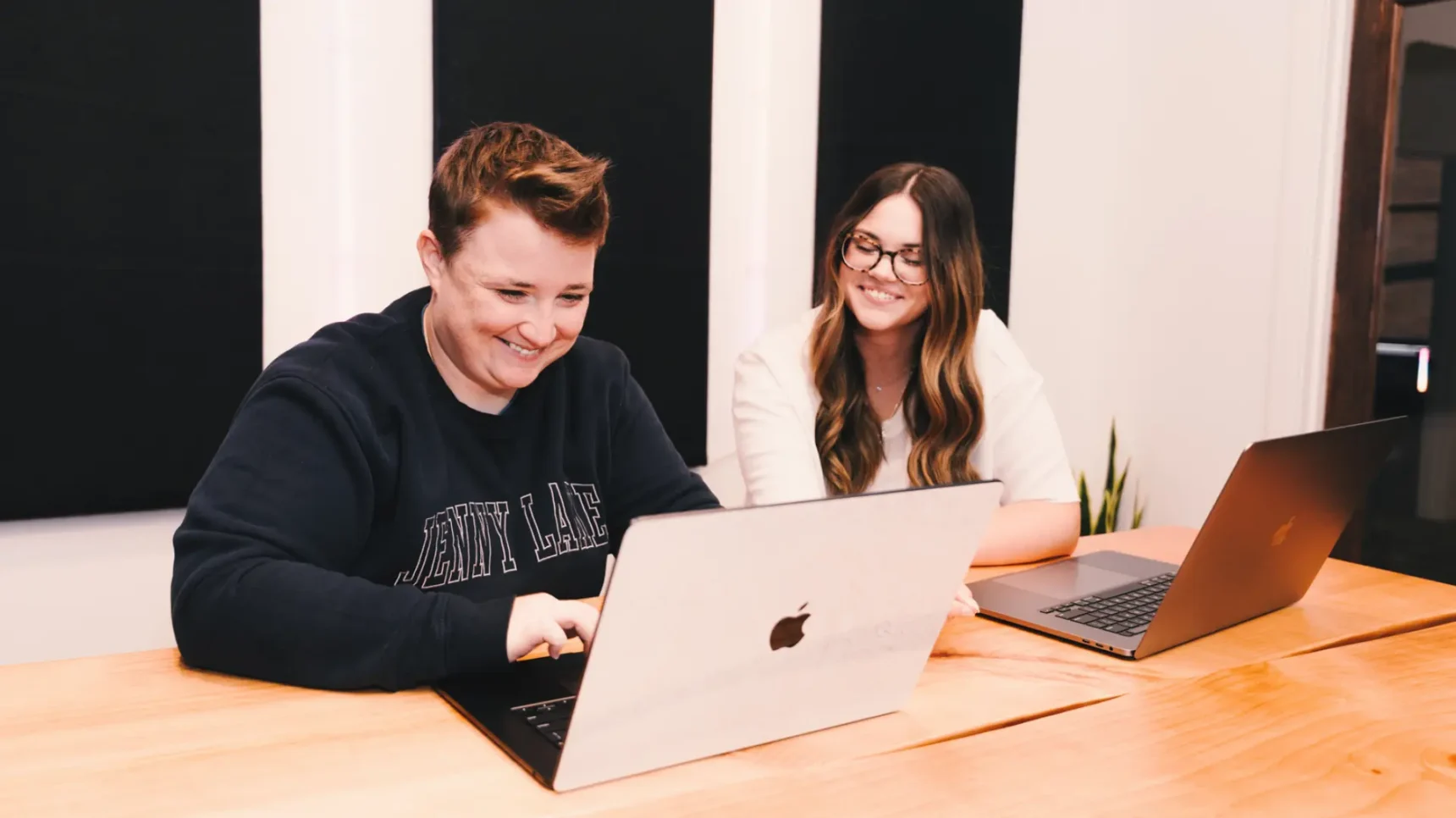 Two individuals are seated at a wooden table, each with a laptop in front of them. The person on the left is wearing a navy sweatshirt and smiling while looking at their laptop. The person on the right is wearing a white shirt and glasses, also looking at their laptop and smiling.