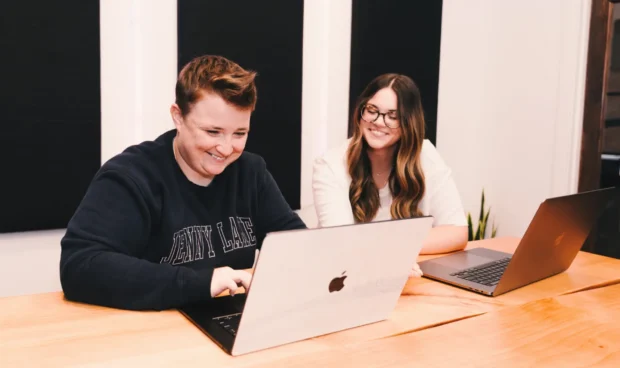 Two individuals are seated at a wooden table, each with a laptop in front of them. The person on the left is wearing a navy sweatshirt and smiling while looking at their laptop. The person on the right is wearing a white shirt and glasses, also looking at their laptop and smiling.