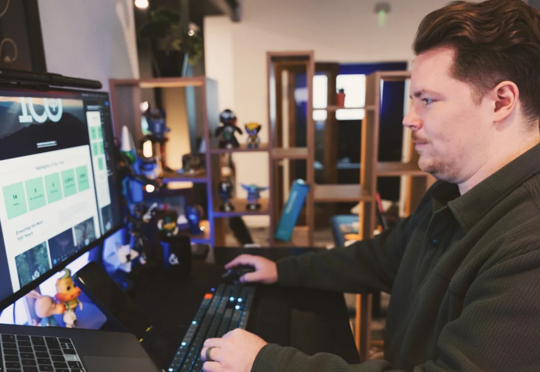 A person in a dark green shirt works at a desk with multiple monitors and a laptop, using a keyboard and mouse. Shelving with decorative items is visible in the background.