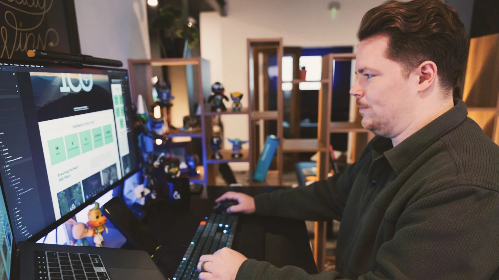 A person in a dark green shirt works at a desk with multiple monitors and a laptop, using a keyboard and mouse. Shelving with decorative items is visible in the background.