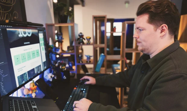 A person in a dark green shirt works at a desk with multiple monitors and a laptop, using a keyboard and mouse. Shelving with decorative items is visible in the background.