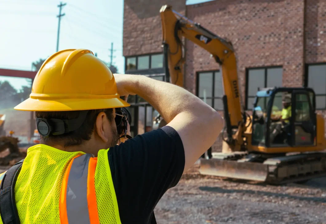 A person wearing a yellow hard hat and a bright yellow safety vest with reflective stripes is taking a photo on a construction site. In the background, an excavator is visible in front of a brick building under construction.