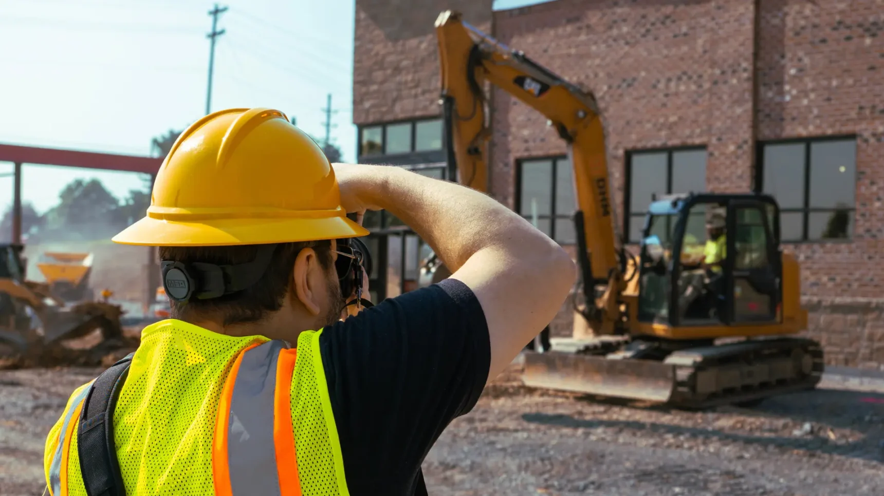 A person wearing a yellow hard hat and a bright yellow safety vest with reflective stripes is taking a photo on a construction site. In the background, an excavator is visible in front of a brick building under construction.