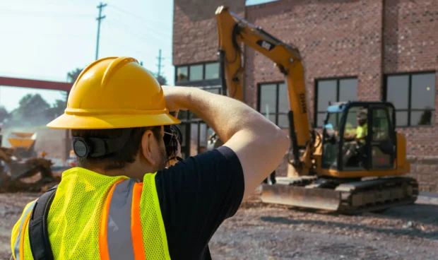 A person wearing a yellow hard hat and a bright yellow safety vest with reflective stripes is taking a photo on a construction site. In the background, an excavator is visible in front of a brick building under construction.