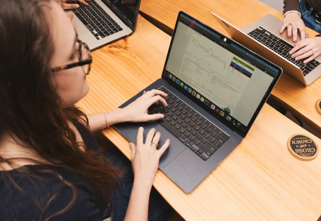 Several individuals working on laptops at a wooden table. The focus is on one person wearing glasses and typing on a silver laptop, with a graph displayed on the screen.