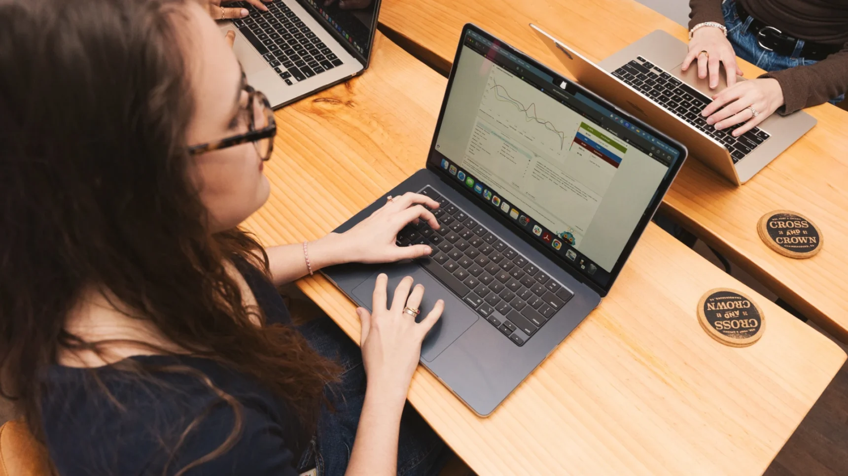 Several individuals working on laptops at a wooden table. The focus is on one person wearing glasses and typing on a silver laptop, with a graph displayed on the screen.
