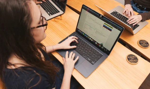 Several individuals working on laptops at a wooden table. The focus is on one person wearing glasses and typing on a silver laptop, with a graph displayed on the screen.