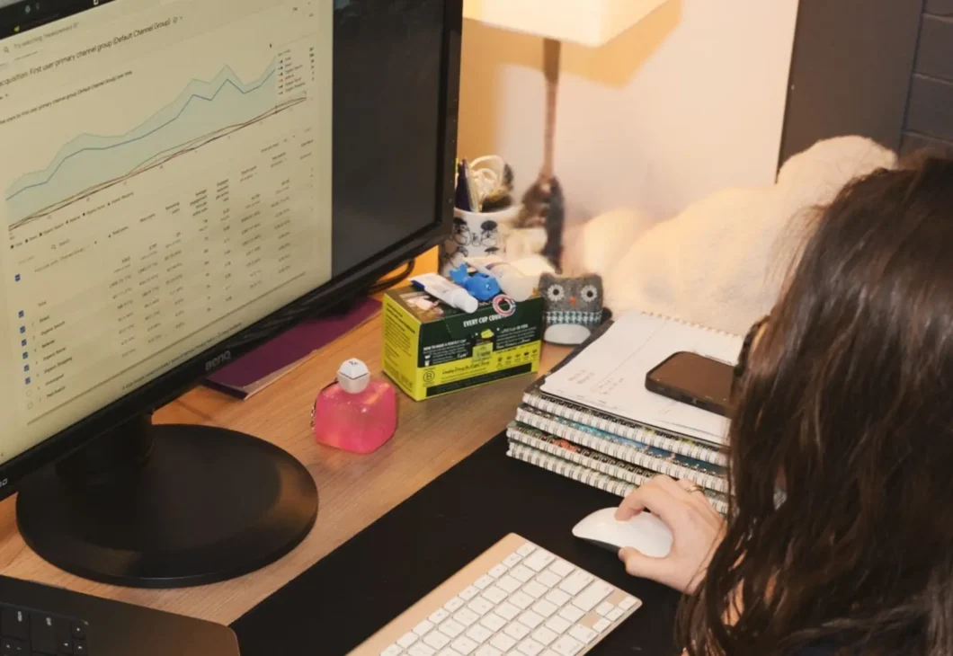 A person sits at a desk with a laptop, a monitor displaying charts, and a keyboard and mouse.