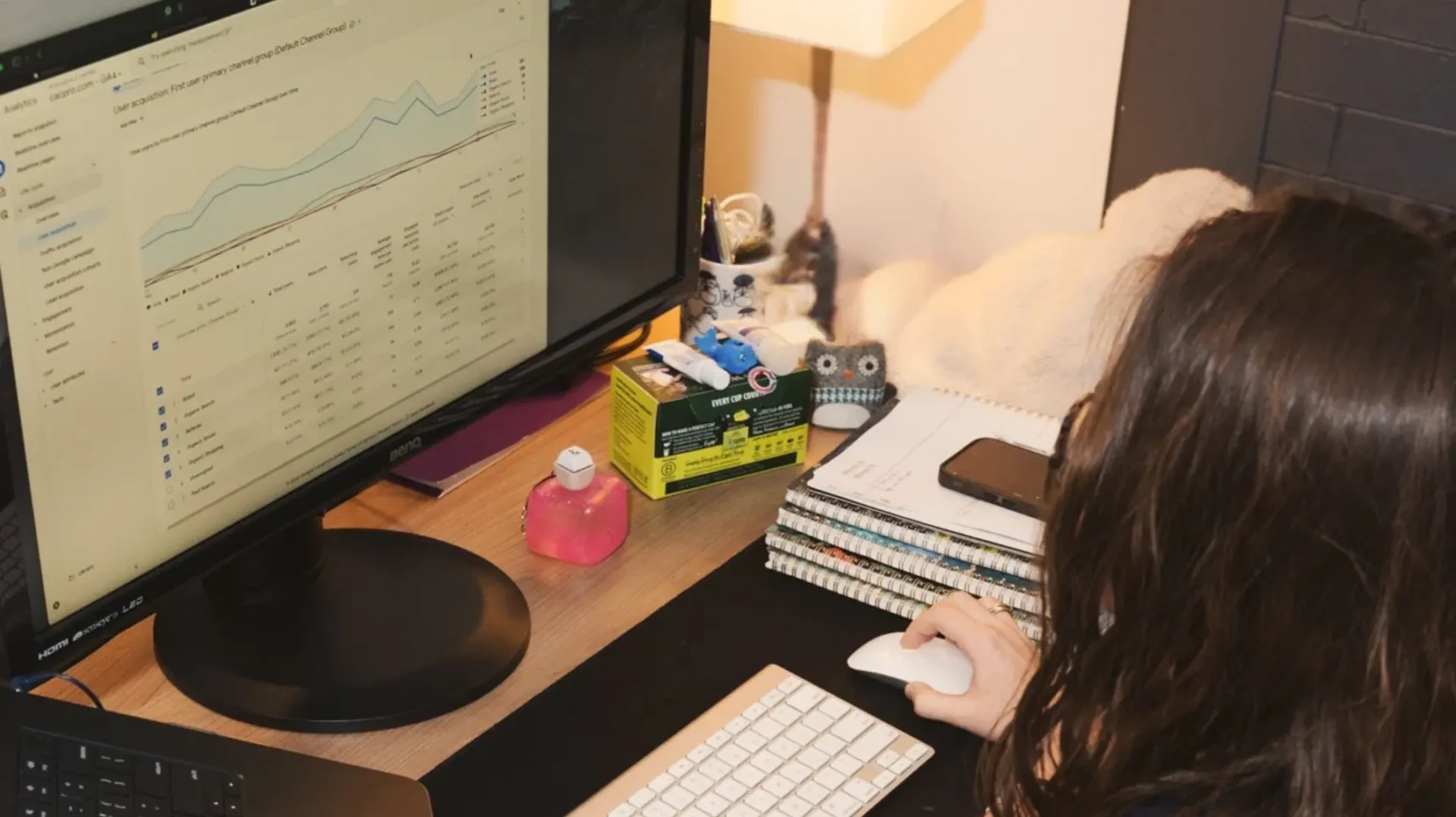 A person sits at a desk with a laptop, a monitor displaying charts, and a keyboard and mouse.
