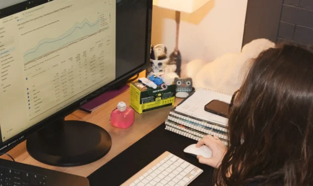 A person sits at a desk with a laptop, a monitor displaying charts, and a keyboard and mouse.