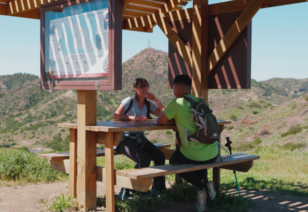 Two hikers sit at a picnic table under a wooden shelter, looking at a map, with rolling hills in the background.