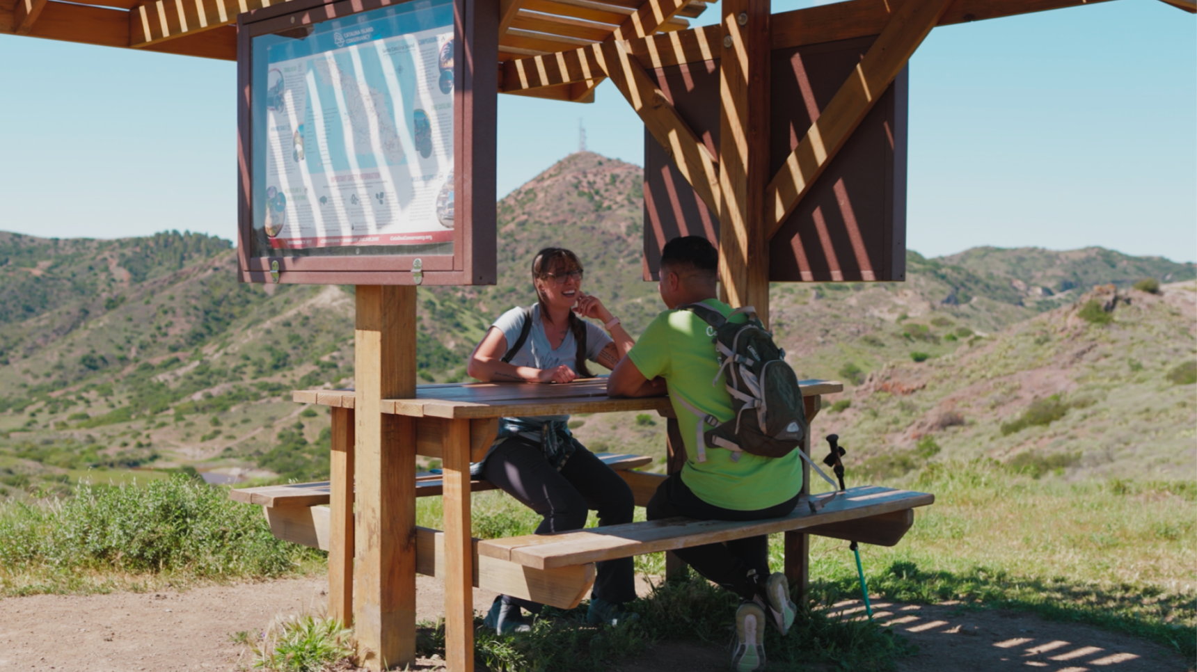 Two hikers sit at a picnic table under a wooden shelter, looking at a map, with rolling hills in the background.