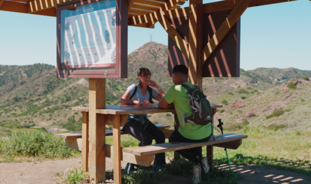 Two hikers sit at a picnic table under a wooden shelter, looking at a map, with rolling hills in the background.