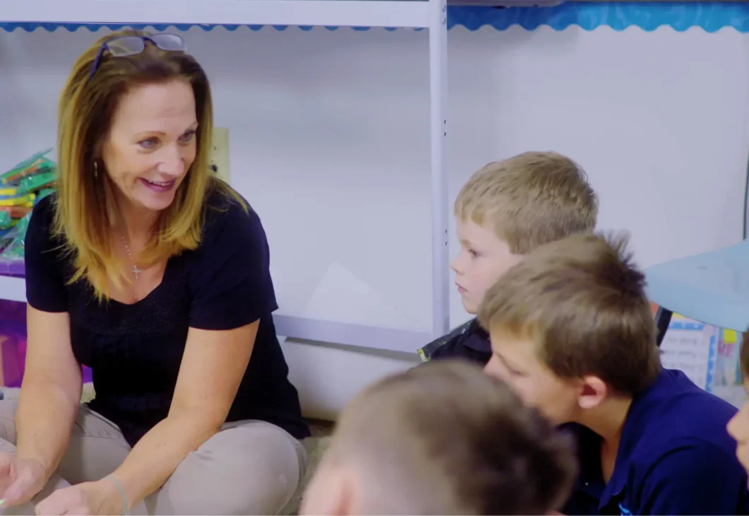 A woman in a black shirt and tan pants sits on the floor, talking to a group of children in a classroom.