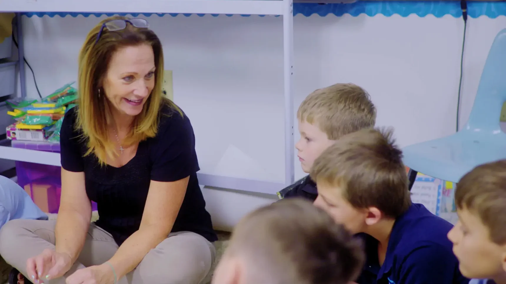A woman in a black shirt and tan pants sits on the floor, talking to a group of children in a classroom.