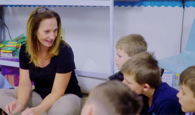 A woman in a black shirt and tan pants sits on the floor, talking to a group of children in a classroom.