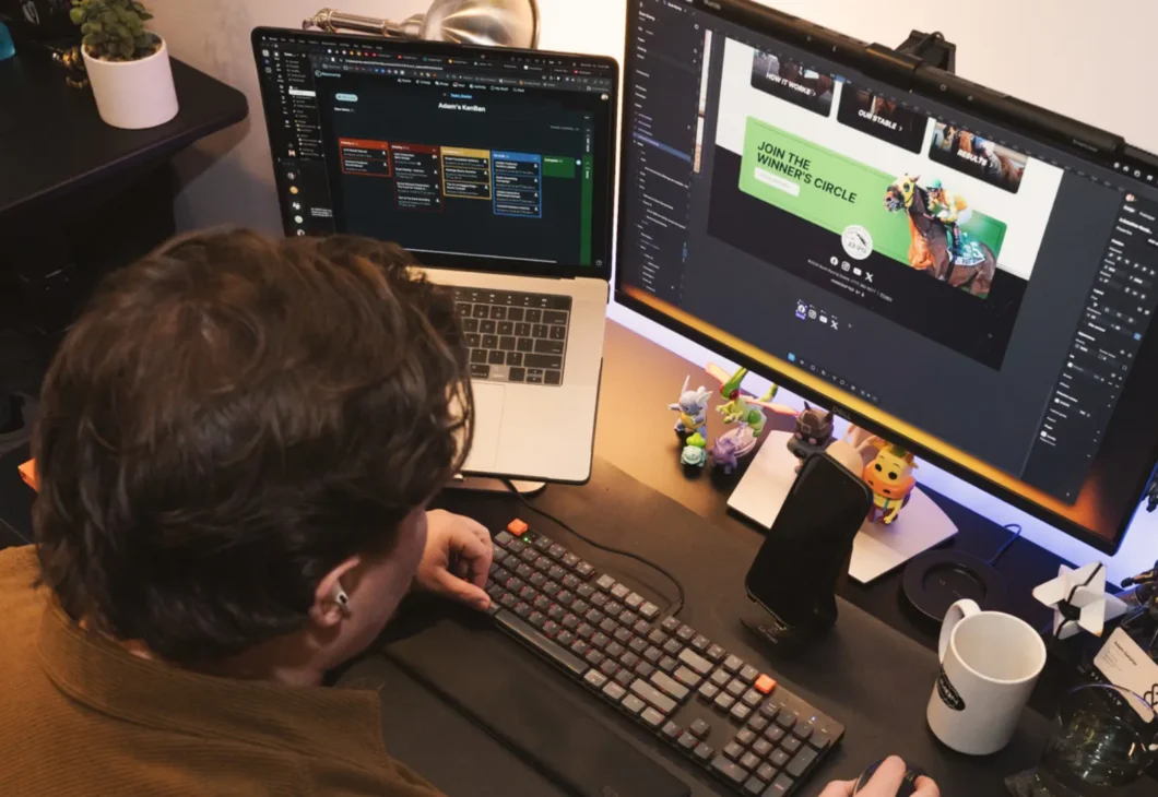 A person wearing a brown sweater sits at a desk with two computer monitors, a laptop, and a keyboard, surrounded by various figurines and desk accessories.