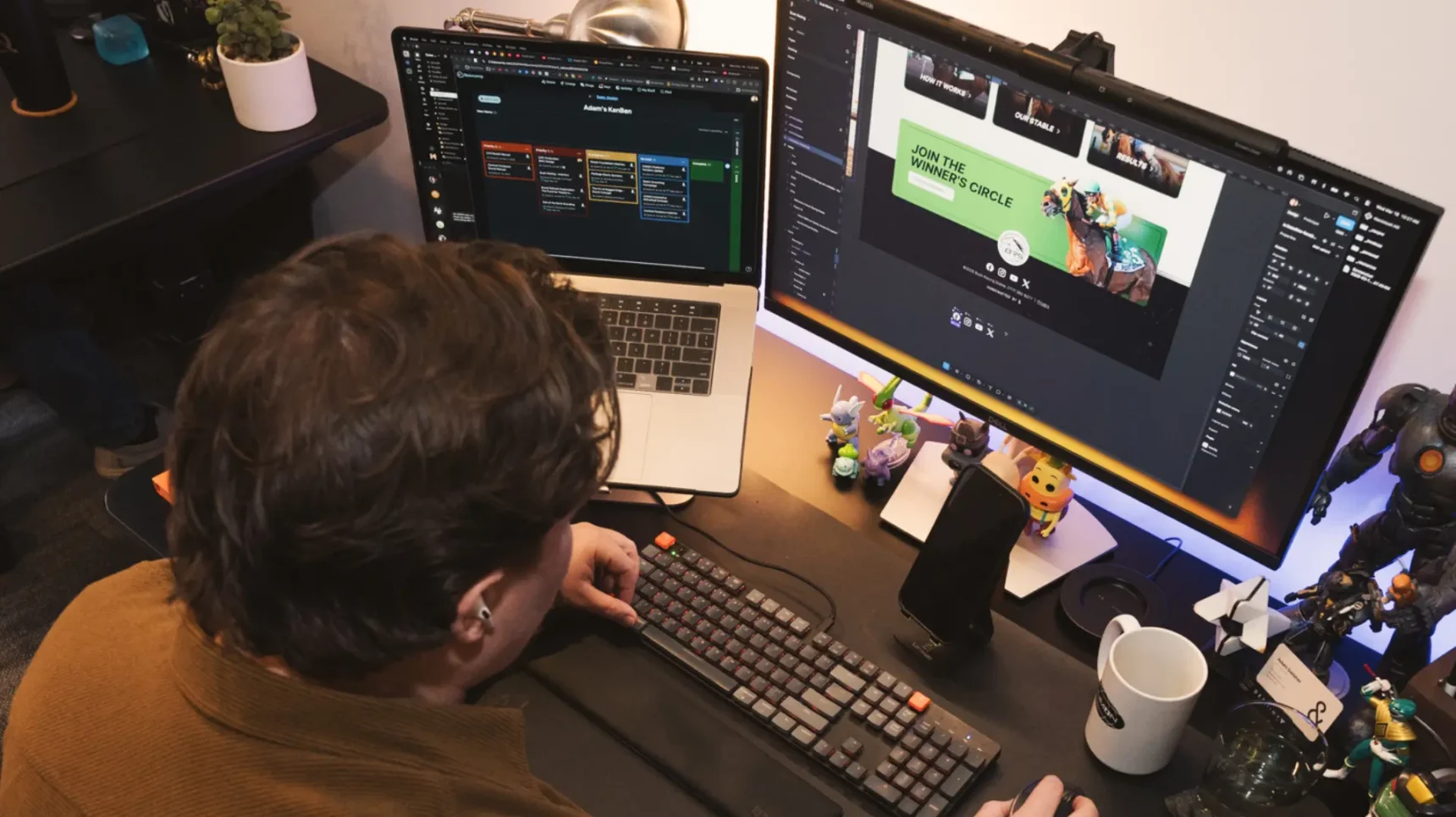 A person wearing a brown sweater sits at a desk with two computer monitors, a laptop, and a keyboard, surrounded by various figurines and desk accessories.