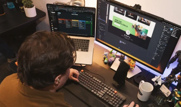 A person wearing a brown sweater sits at a desk with two computer monitors, a laptop, and a keyboard, surrounded by various figurines and desk accessories.