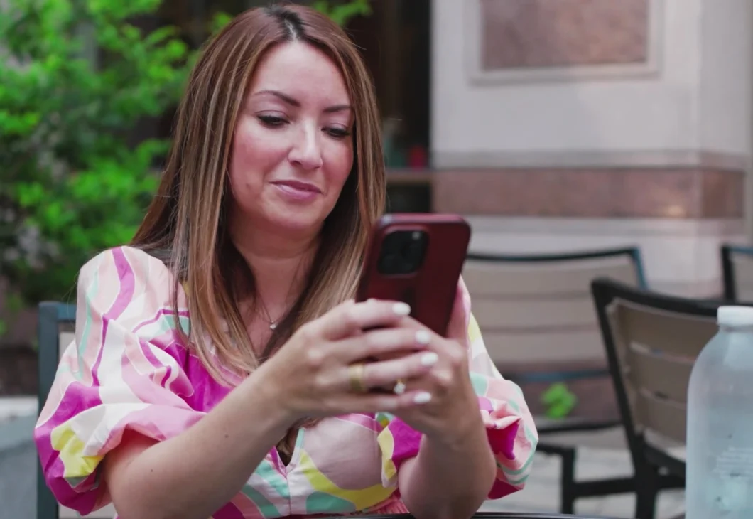 A person in a colorful dress holds a phone and looks at it while sitting at a table outdoors. A bottle of water is on the table.