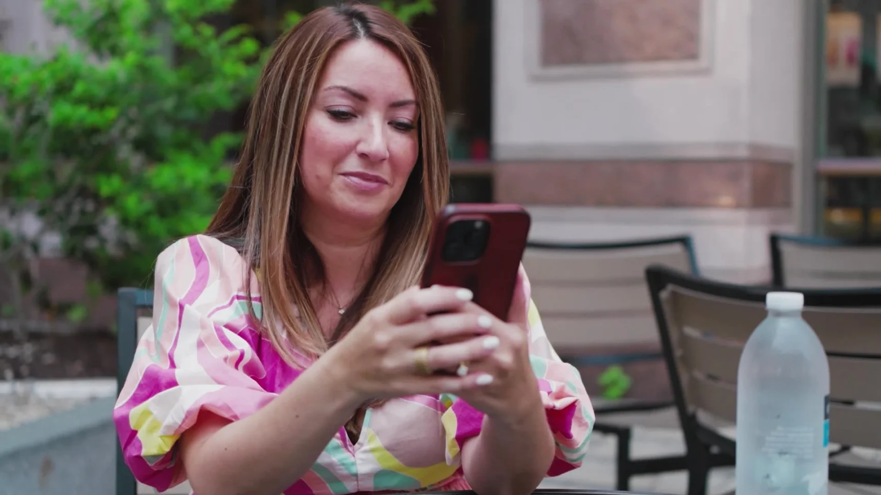 A person in a colorful dress holds a phone and looks at it while sitting at a table outdoors. A bottle of water is on the table.