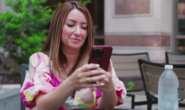 A person in a colorful dress holds a phone and looks at it while sitting at a table outdoors. A bottle of water is on the table.