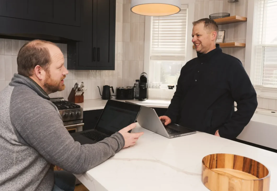Two people in a kitchen, one seated at a counter with a laptop and the other standing nearby with a laptop, both appear to be conversing.