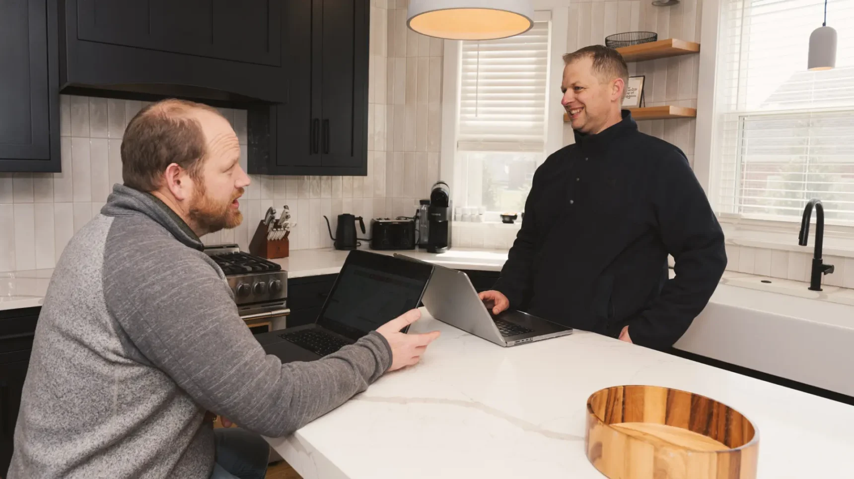 Two people in a kitchen, one seated at a counter with a laptop and the other standing nearby with a laptop, both appear to be conversing.