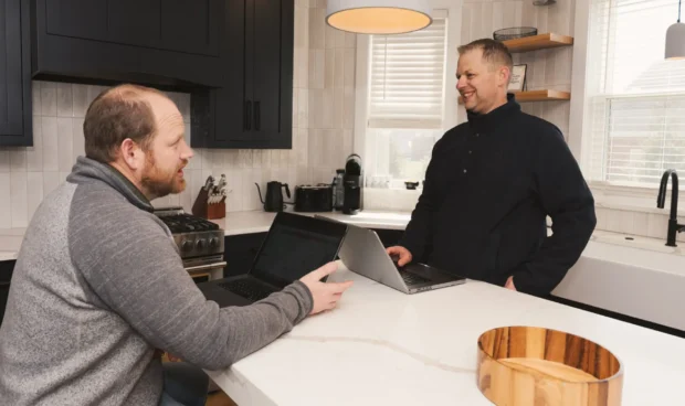 Two people in a kitchen, one seated at a counter with a laptop and the other standing nearby with a laptop, both appear to be conversing.