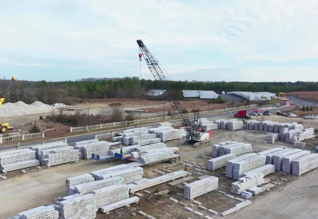 An aerial view of a construction site with stacked concrete slabs, cranes, and trucks under a cloudy sky.