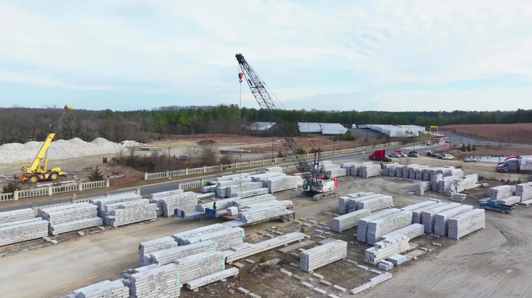An aerial view of a construction site with stacked concrete slabs, cranes, and trucks under a cloudy sky.