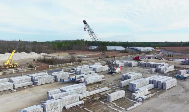 An aerial view of a construction site with stacked concrete slabs, cranes, and trucks under a cloudy sky.