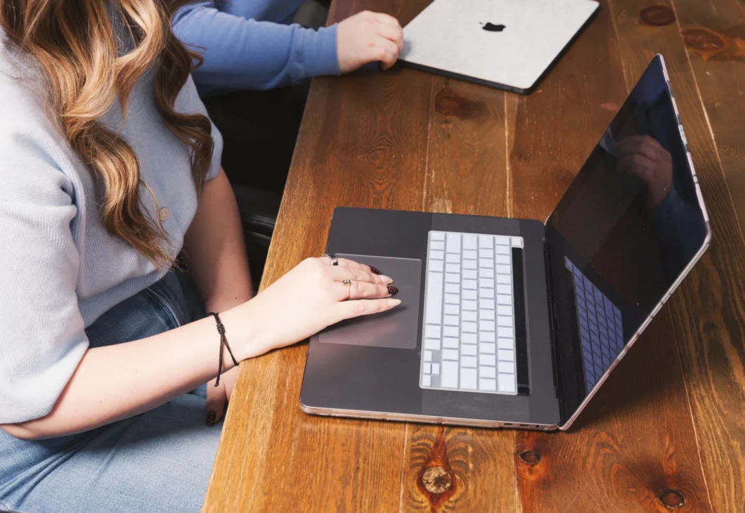 A person's hands are on the keyboard and trackpad of an open laptop computer on a wooden desk. Another laptop is visible in the background.