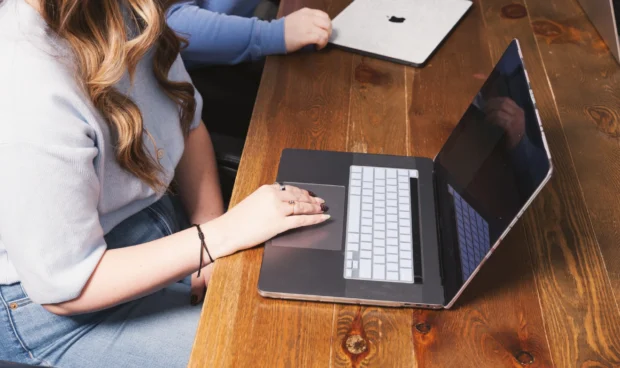 A person's hands are on the keyboard and trackpad of an open laptop computer on a wooden desk. Another laptop is visible in the background.
