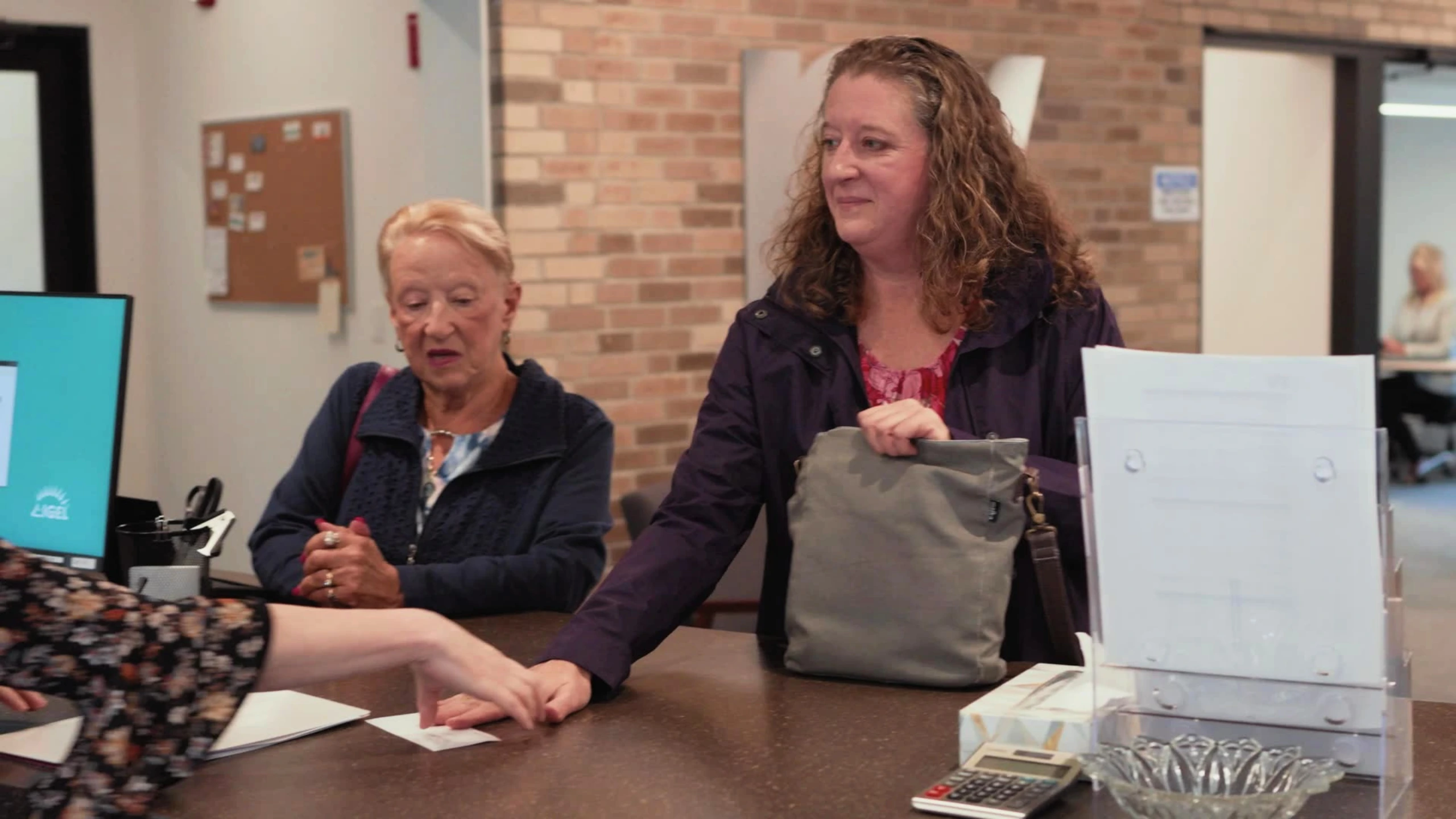 Two people interact at a reception desk with a computer monitor and paperwork visible.