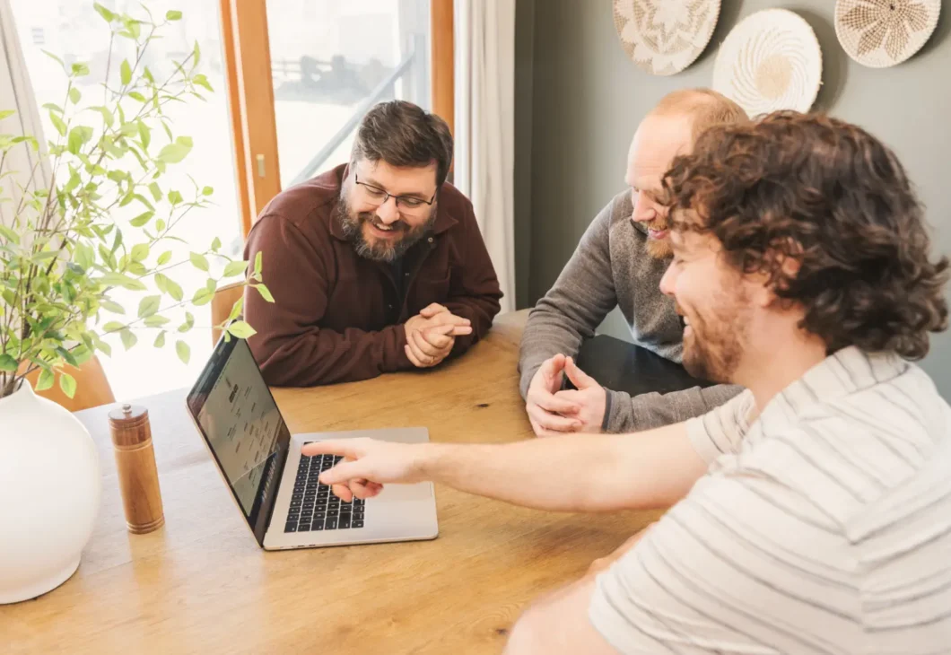 Three individuals gathered around a laptop on a wooden table, with one person pointing at the screen.