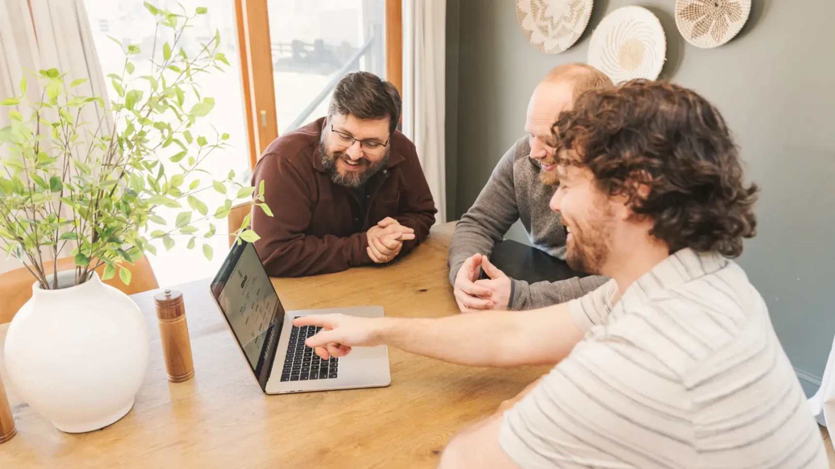 Three individuals gathered around a laptop on a wooden table, with one person pointing at the screen.