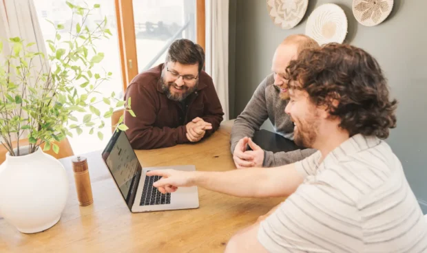 Three individuals gathered around a laptop on a wooden table, with one person pointing at the screen.