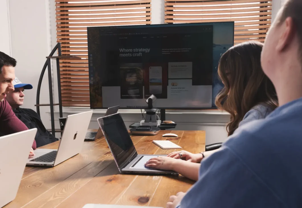 People collaborating at a desk with laptops in an office meeting, one person writing on a notepad while another views a monitor behind them, with a coffee mug on the table.